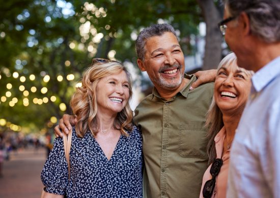 Group of happy senior friends sharing a moment outdoor while embrace. Older men and laughing women chatting together during a walk. Close up face of cheerful retirees enjoying time in a lively city street.